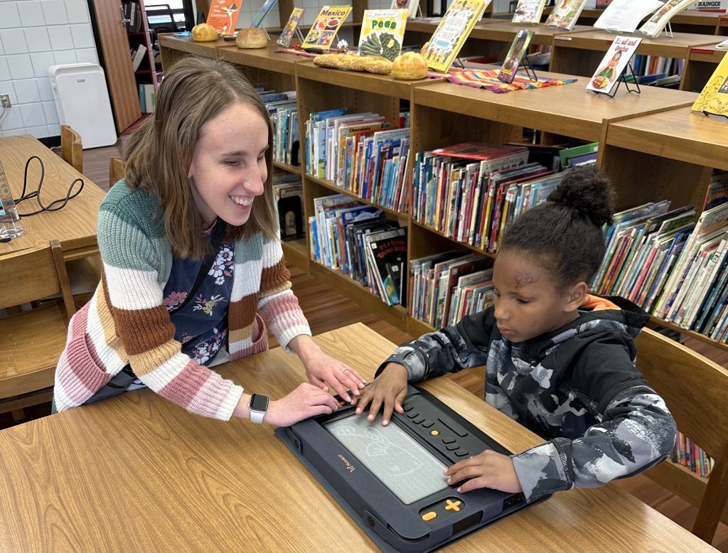 A woman in a striped sweater helps a young boy use the Monarch on a table in a school library.