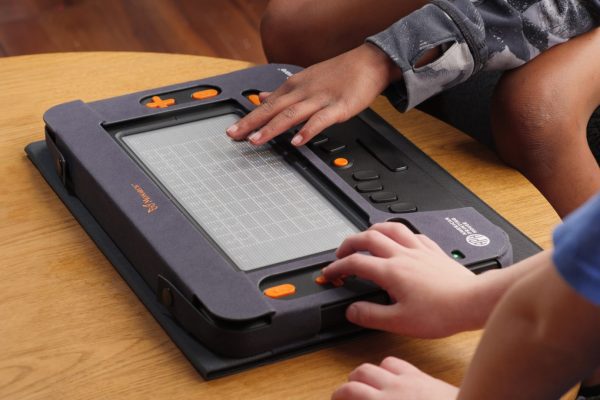 A top-down view of two boys playing a game of chess on the Monarch. The display is rendering a tactile chess board, and each boy has one hand on the device, feeling the display and the directional keys.