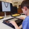 A young student in a library using a Monarch connected to a visual display. As he explores a tactile parabola graphed on the Monarch display using the KeyMath app, the same image is mirrored on a monitor above.
