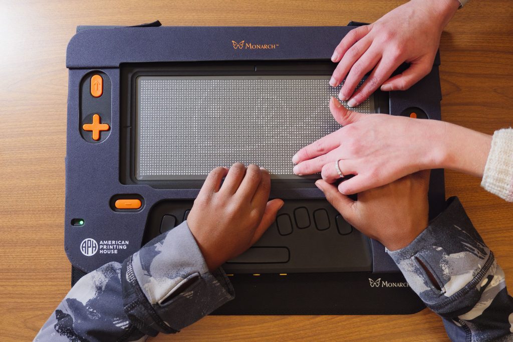 Two adult hands guide a child's hands across a tactile graphic of a fish displayed on the Monarch's refreshable braille display.