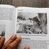 A hand holding open a copy of Tools for TVIs to a page showing a student working on a Perkins brailler with adult support.