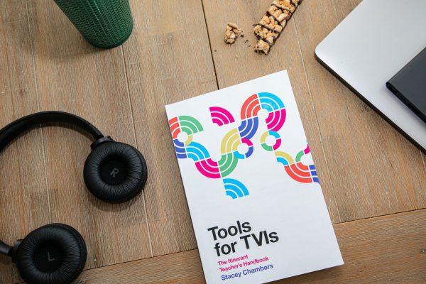 Printed copy of Tools for TVIs on a busy wooden workspace surrounded by a pair of headphones, a mug, a laptop, and a partially eaten granola bar.