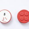 Close up comparison of the front and back of a red circular UEB Reach and Match number tile showing the number 5 in braille and print. The back of the piece is textured.