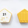Close up comparison of the front and back of a yellow hexagonal UEB Reach and Match number tile showing the number 21 in braille and print. The back of the piece is textured.