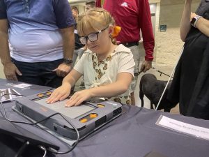 A young girl wearing glasses and an outfit with Monarch butterflies on it touches a tactile graphic on the Monarch’s multiline refreshable braille display as several adults and a black guide dog stand behind her.