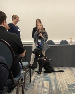 A young woman with a black guide dog seated in front of her stands in front of a table and speaks to a crowd while holding a microphone. Two other women stand behind and beside her.
