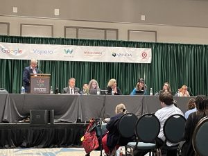 APH President Craig Meador stands behind a podium on a stage, and to his left, several people are seated at a table with microphones on it. A seated crowd is facing the stage.
