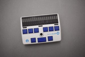 The Refreshabraille 18. The rectangular plastic case is silver and black with slightly rounded sides. An eighteen-cell, eight-dot refreshable braille display is at the top of the device, with a round cursor routing button below each cell and three display advance bars below the display. Nine square blue braille entry keys are arranged six over three beneath the display advance bars and the joystick, which is located in middle of the device. The APH logo and
