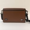 Front view of a closed turtleback case on a white background. The strap sits to the right of the device, and the brown cover has logos from HumanWare and APH