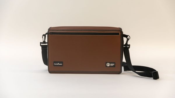 Front view of a closed turtleback case on a white background. The strap sits to the right of the device, and the brown cover has logos from HumanWare and APH