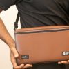A man wearing a Mantis across his body in the Turtleback case and holding the closed device with both hands. The front of the brown case contains logos for HumanWare and APH.