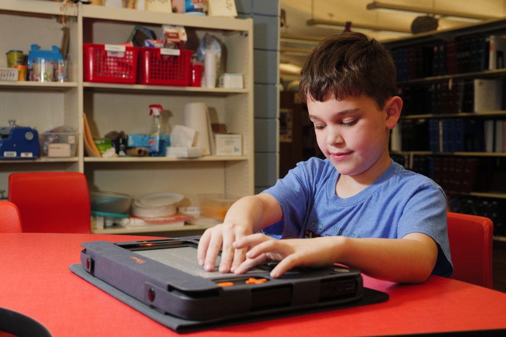 A young boy reads with both hands on the Monarch's multiline refreshable braille display on a red table in a classroom.