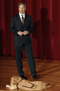 Mike May wears a suit as a speaks in front of a red backdrop with a guide dog lying at his feet.