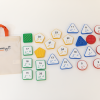 All the tiles contained in the Reach & Match Number Tiles (UEB) kit laid out next to a canvas Reach & Match storage bag. The tiles come in red circles, blue triangles, yellow hexagons, and green squares. The front of the tiles contain a printed number with the corresponding UEB braille label, and the backs have various texture patterns.