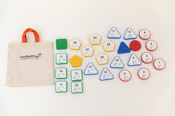 All the tiles contained in the Reach & Match Number Tiles (UEB) kit laid out next to a canvas Reach & Match storage bag. The tiles come in red circles, blue triangles, yellow hexagons, and green squares. The front of the tiles contain a printed number with the corresponding UEB braille label, and the backs have various texture patterns.