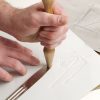 Two hands creating a tactile graphic of shapes and lines using the included hammer and tong tool on white paper.
