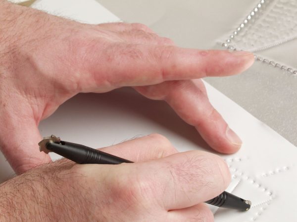 One handing holding a white piece of paper steady while the other hand, holding the tactile graphics kit roller, creates a tactile line.