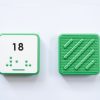 Front and back of green square Reach & Match Number tile showing the number 18 in Nemeth braille and print. The back side is textured.