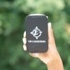 A hand holding an upright VR Cardinal device with a closed case against green foliage in the background. A VR Cardinal logo along with the words “VR CARDINAL” are printed in white on the black case.