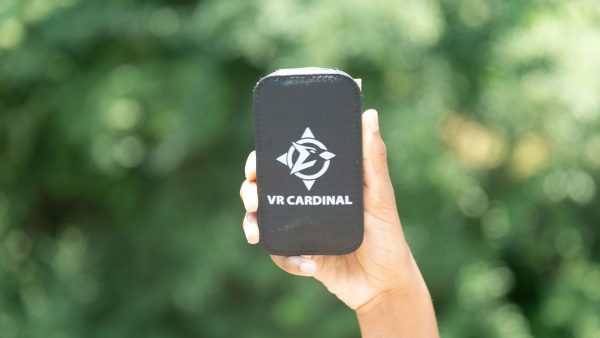 A hand holding an upright VR Cardinal device with a closed case against green foliage in the background. A VR Cardinal logo along with the words “VR CARDINAL” are printed in white on the black case.