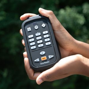Two hands cradling an open VR Cardinal device against a background of dark green foliage. On the face of the device are various buttons, and along the bottom underneath the play button, “VictorReader” is printed in white.