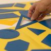 One hand holds an outer triangle stencil up. The inner triangle stencil is adhered to the yellow folder on a white background.