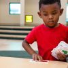 A preschool aged boy in a red shirt holding several Memory Shapes puzzle pieces in one hand and picking up another piece with his other hand.