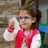 A preschool girl in a white and pink letterman-style jacket and purple glasses holds a crescent shape puzzle piece up to her face to look at it.