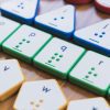 Four rows of Reach & Match Alphabet Tiles on a wooden floor. The row of square green tiles are in focus with blue triangles and red circles above and a row of yellow hexagonal tiles beneath.