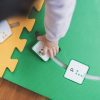 A child's hand inserts a green square alphabet tile into the green mat on a wooden floor.