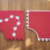 Two red Reach & Match mats side by side demonstrating the differences between the textured side and the side with circular cut-outs for tiles and a traceable path.