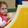 A little girl in purple glasses holding a yellow Reach and Match tile up to her eyes while making an inquisitive facial expression.
