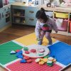A little girl plays with Reach and Match tiles assembled in a square with their textured sides visible. The tile carrying case is placed in the center of the mats.