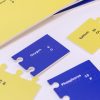 Close up of a blue oxygen piece and a yellow calcium piece with other pieces scattered around out of focus. Each piece has the name of the element, the atomic number, and the atomic symbol in both print and braille.