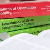 The red hardback Foundations of O&M volume II on top of a stack of green Foundations books. The title and editors' names are printed on the spine in white text.