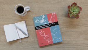 An overhead shot of a copy of Accessible Science sitting on a wooden surface next to a mug of black coffee, a notebook and pen, and a green and red succulent.