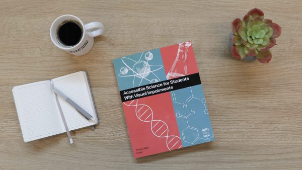 An overhead shot of a copy of Accessible Science sitting on a wooden surface next to a mug of black coffee, a notebook and pen, and a green and red succulent.