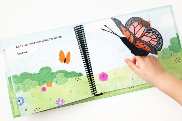The hand of a young child is playing with a Monarch butterfly manipulative included in On the Way to Literacy: The Caterpillar. The book is opened to a page with bright and colorful illustrations of a butterfly flying over a field.