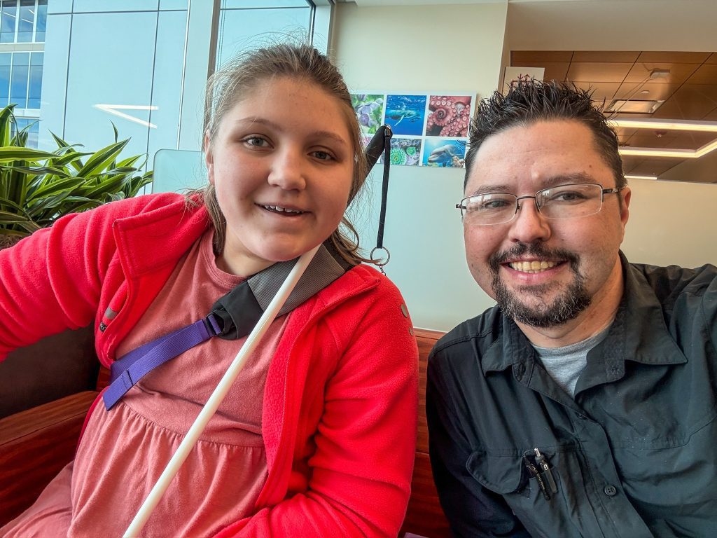 On the left Isabelle, holding her cane, smiles at the camera. To her right sits her dad Rob, also smiling at the camera.