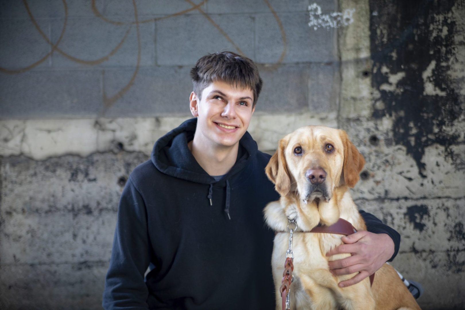 A young man in a black sweatshirt smiles at the camera. His arm is around the golden guide dog sitting on his right.