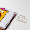 An open page of My Nose with the character illustration on the right. The main character has a red bag where their nose should be. On the right is large-print text with orange embossed braille. The book is spiral bound.