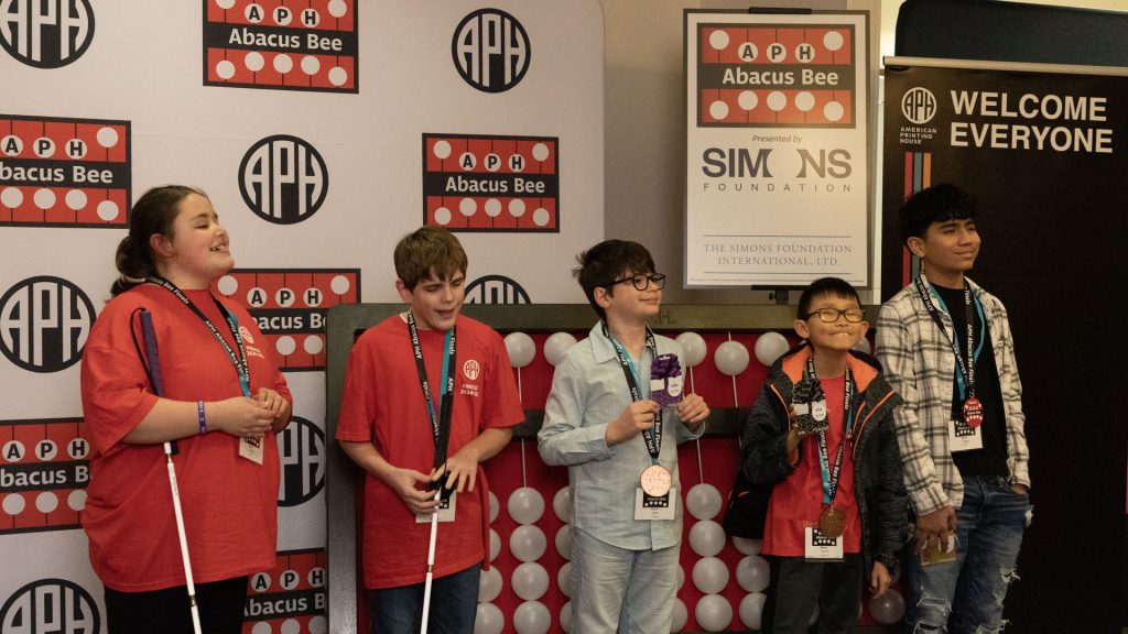 5 students of various ages and races standing in front of a giant abacus and different signage with APH, Abacus Bee, and Simons Foundation logos.