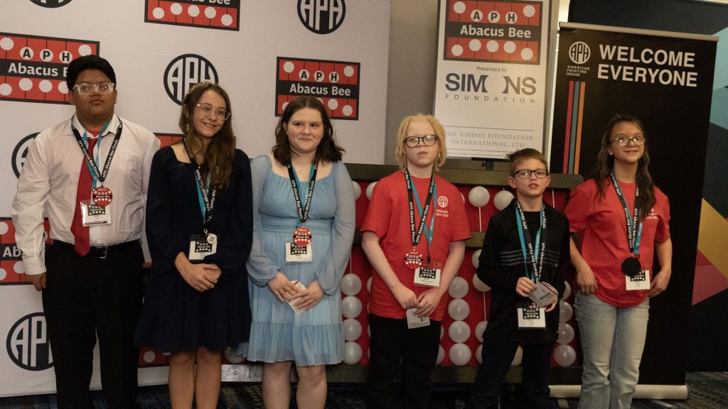 Six students of various ages and races standing in front of a giant abacus and different signage with APH, Abacus Bee, and Simons Foundation logos.