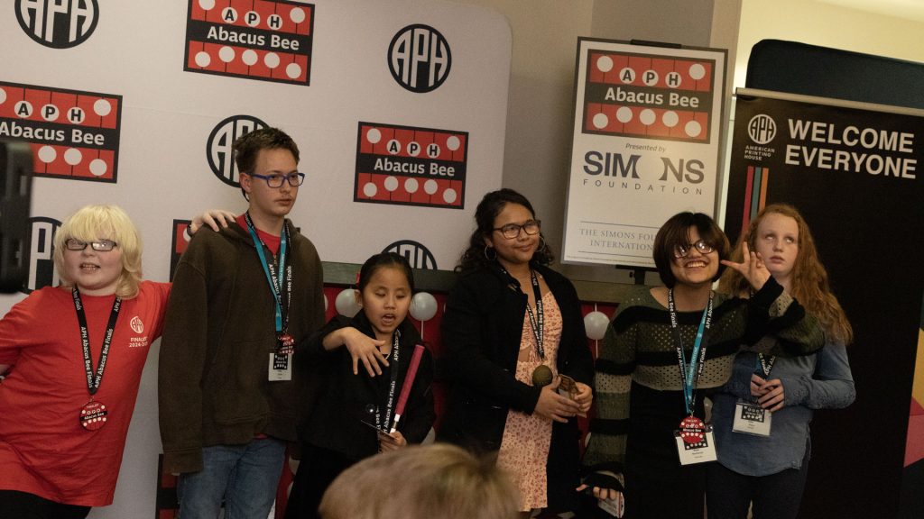 6 students of various ages and races standing in front of a giant abacus and different signage with APH, Abacus Bee, and Simons Foundation logos.