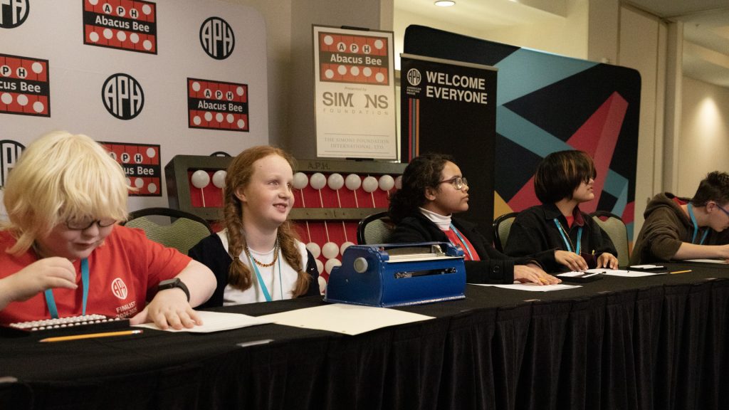 5 students sitting along a table competing in an Abacus Bee competition. Various signage is in the background with APH, Abacus Bee, and Simons Foundation logos along with a giant Abacus.