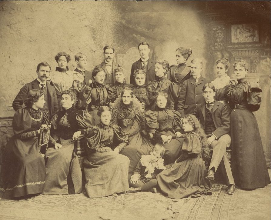 The First Class of the Wright Humason School for the deaf, NYC. Helen Keller with Anne Sullivan Macy in a group photograph of the class. Keller is at the far left of those seated in the first row, she holds Macy's left hand with her right hand. She is wearing a long dress with a fitted waist and wide skirt. The dress has gigot (puff) sleeves and a ruched bodice and a high ruched neckline. Her hair is curled at the bottom to frame her face and head. Macy is seated next to her. Macy wears a dress in the same style, but with a taller collar.
