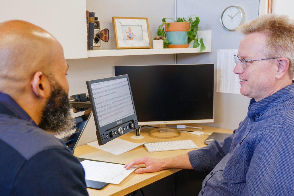 two men talking at a desk with a laptop