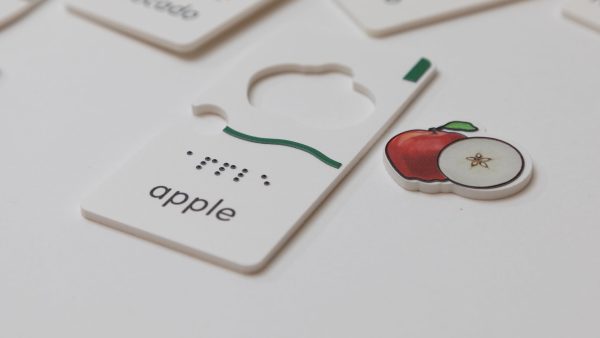 A close-up of the apple puzzle piece that says "apple" in print and braille with the removable apple graphic positioned next to the piece. The piece has a wavy tactile indicator as well as a small tactile square in the top right corner.