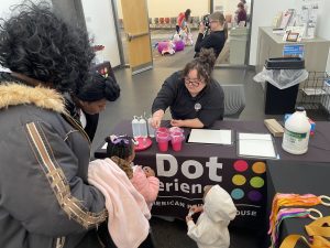 A woman wearing an APH polo leans forward across a table to help two young children with an activity. The table is covered with a black table cloth featuring the Dot Experience logo/