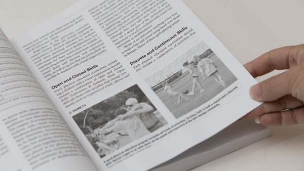 An open page of PE and Sports with a hand holding the bottom right corner of the page. There are two photos, one of a student learning archery from a teacher and another of three girls playing soccer.